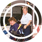 A red rose transit worker pushing a wheelchair patron onto a bus.